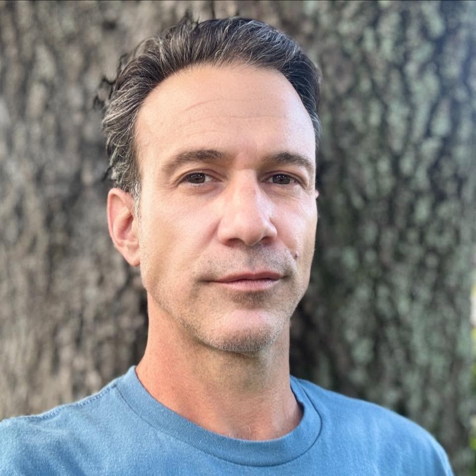 Justin — a man with dark hair and facial hair wearing a blue shirt against a textured stone wall.